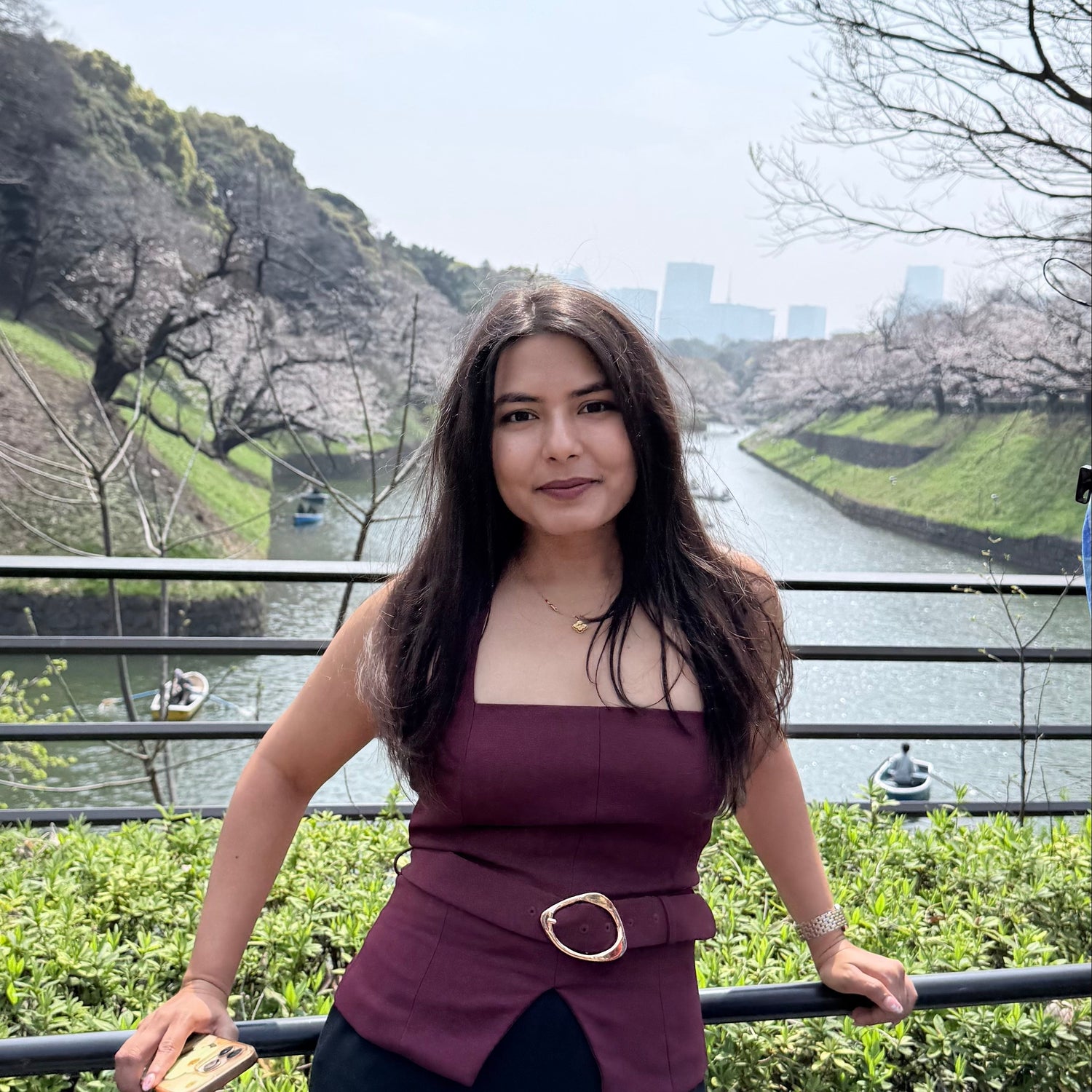 Pankti Bardolia posing outdoors in front of a railing with a river, cherry blossom trees, and distant buildings in the background.